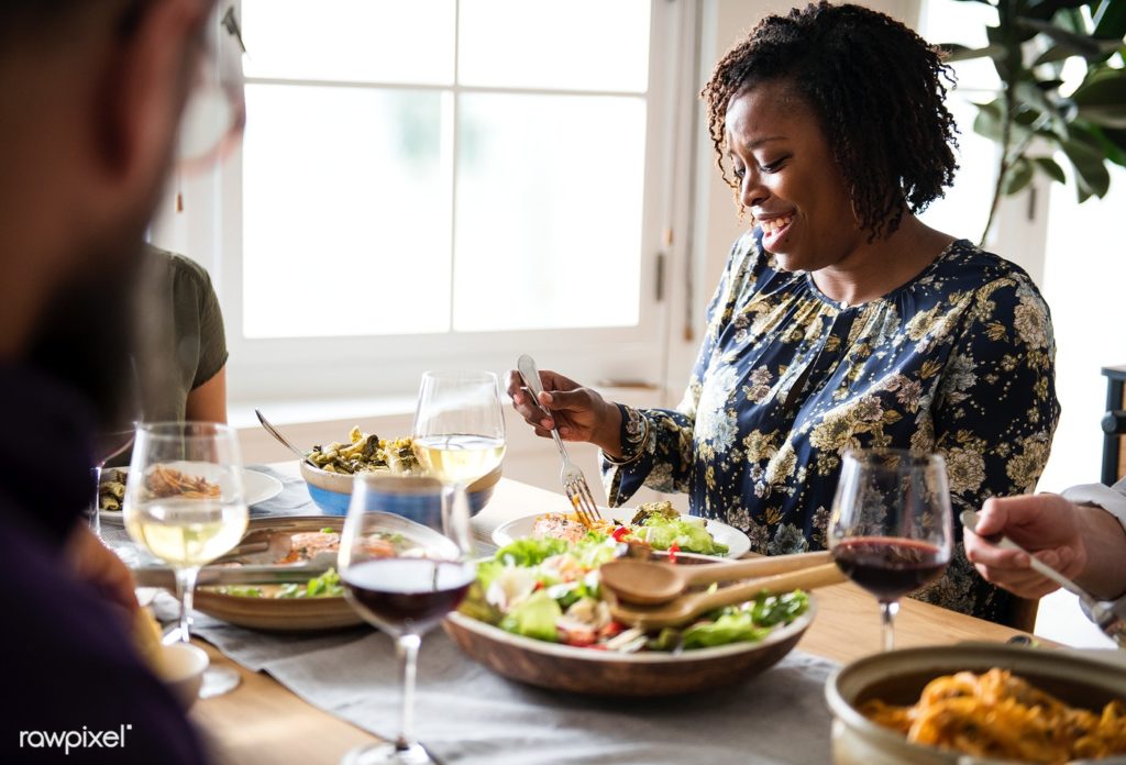 people sitting around a dinner table with the a large salad serving bowl at the center of the table and the focus being on one woman on the right who is picking up food with a fork